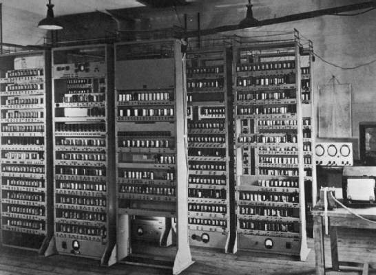 "A black and white photograph shows multiple tall vertical racks of the EDSAC computer system densely populated with vacuum tubes and electronic components. The image captures the large-scale physical architecture of early stored-program computers within a laboratory setting.."